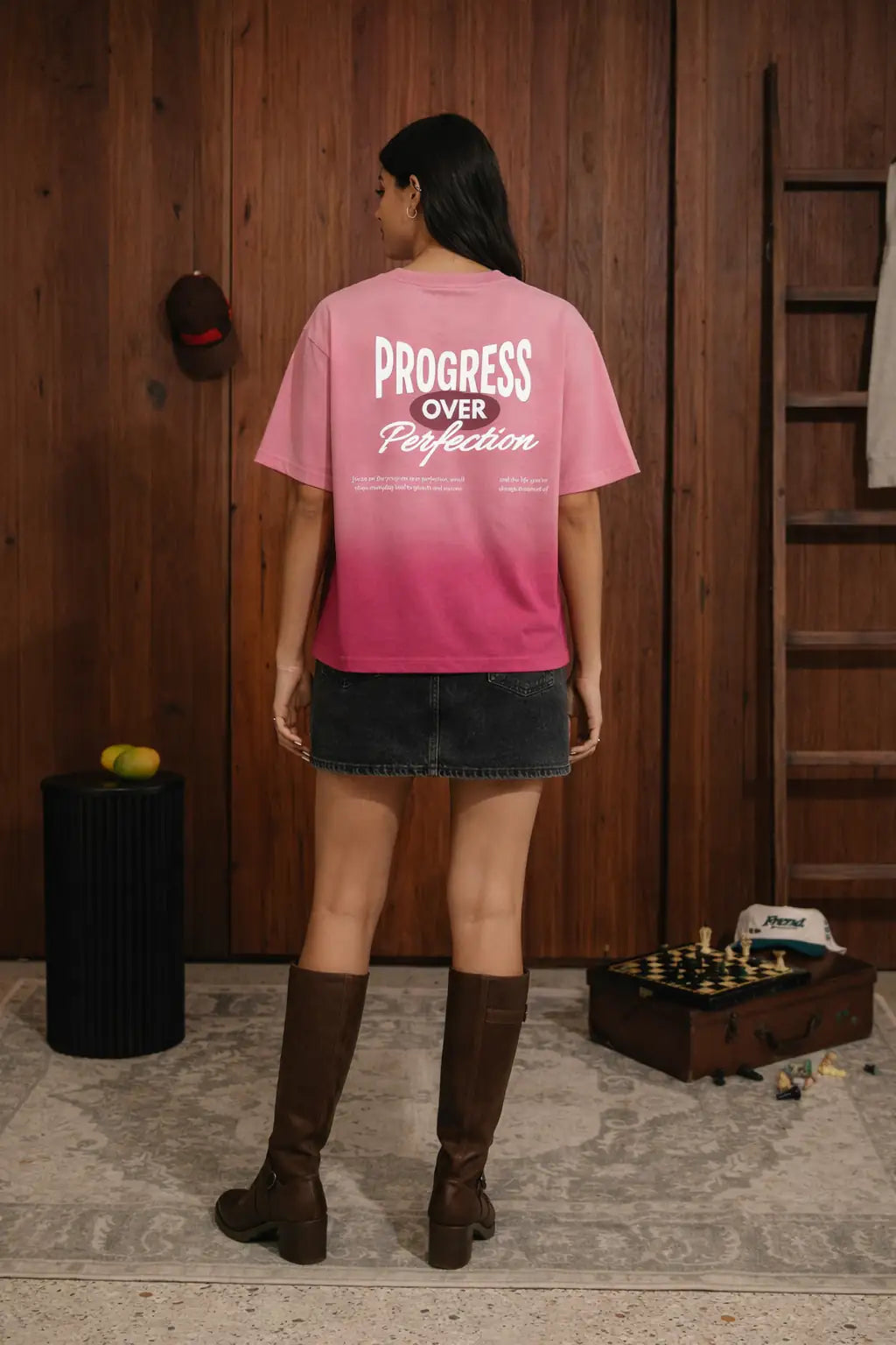 Person wearing a pink t-shirt with 'Progress Over Perfection' text, standing in a room with wooden walls and a chessboard.