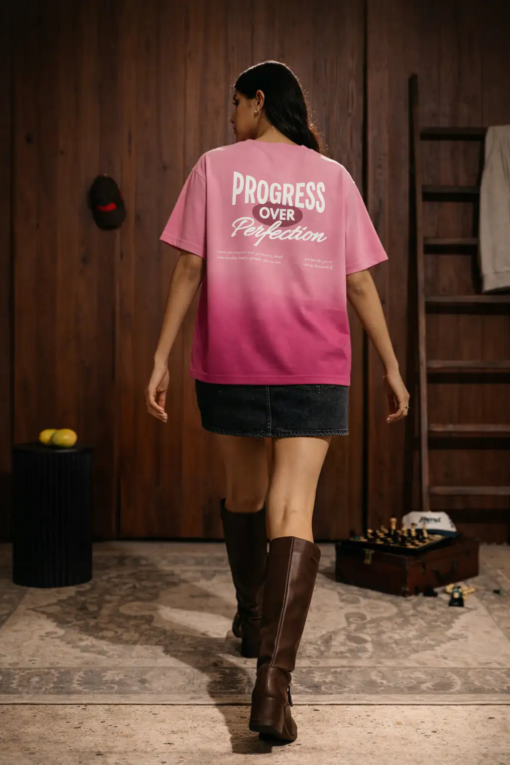 Person wearing a pink t-shirt with 'Progress Over Perfection' text, walking in a room with wooden walls and a rug.